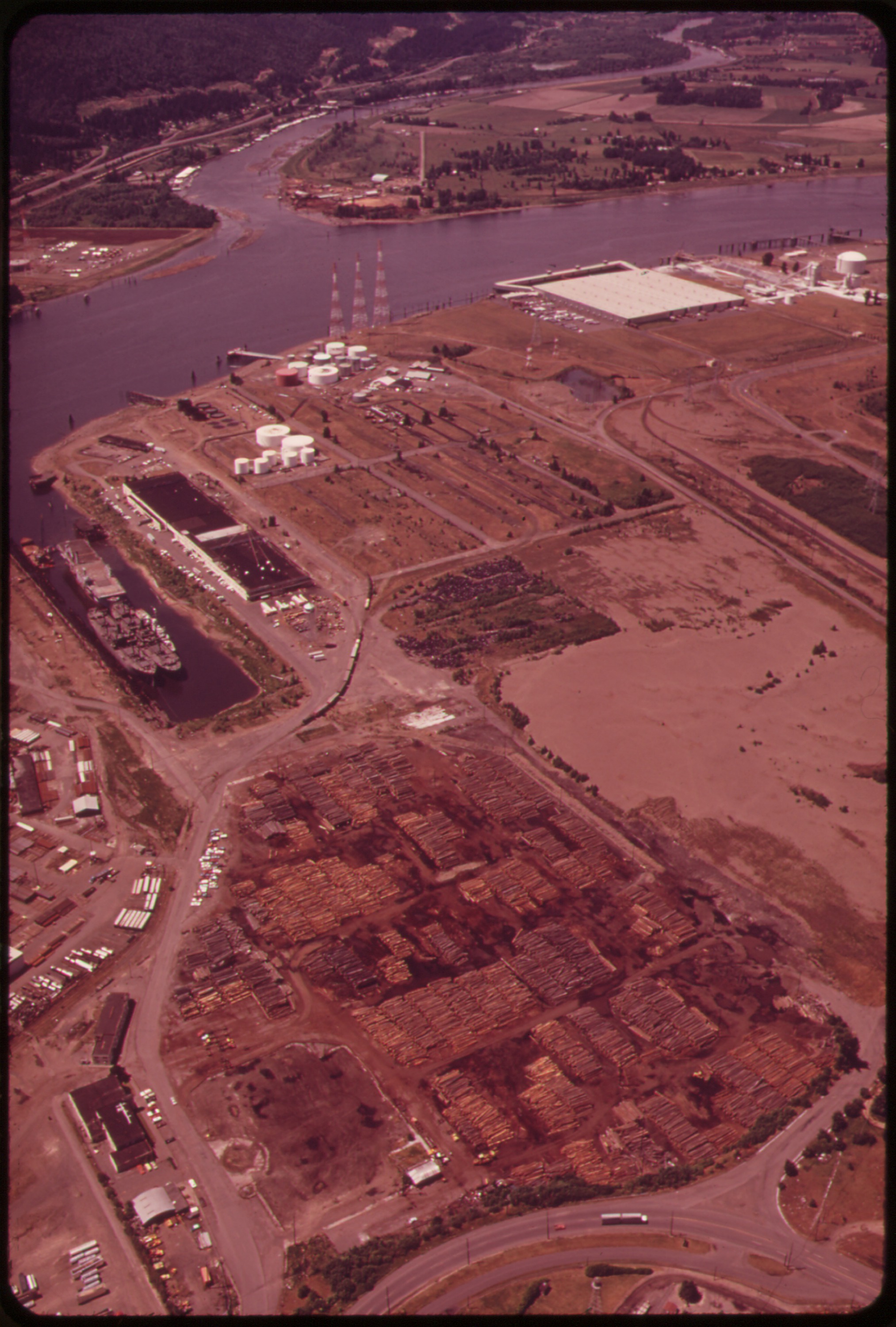 An oblique aerial photograph in saturated reddish tones looking down on an industrial river peninsula. A wide river curves around the land at left, with a moored ship at a small basin, cylindrical storage tanks, a large flat-roofed warehouse, and what appear to be stacks of cut logs arranged in geometric piles across cleared ground. Roads and parking lots wind between the facilities, and forested hills rise in the far distance under a hazy sky.